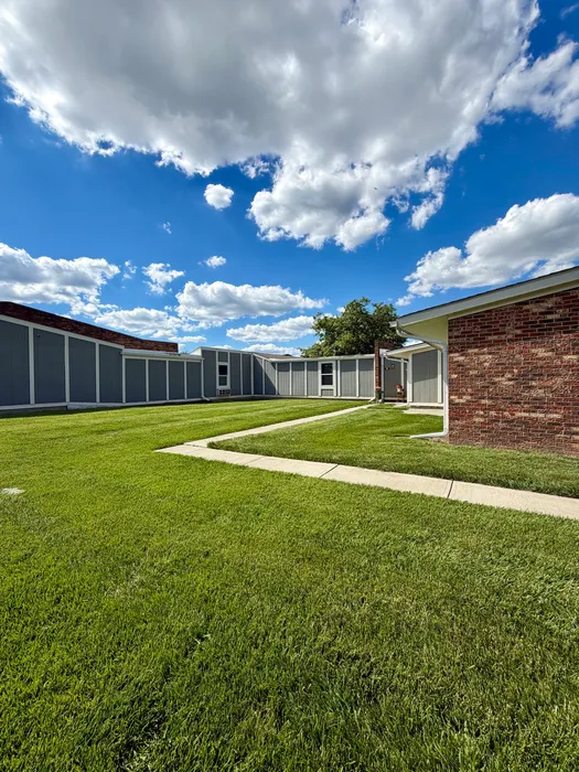 Expansive green lawn with modern buildings under a vibrant blue sky adorned with fluffy clouds.