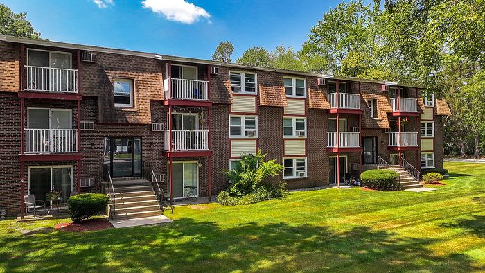 Charming brick apartment building with private balconies and lush green landscaping under a clear blue sky.