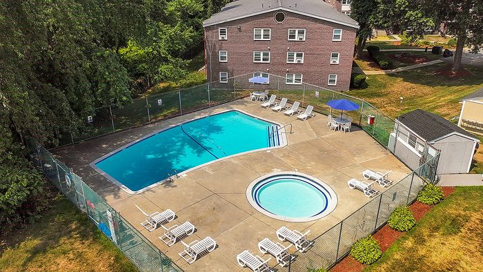 Inviting aerial view of a serene apartment complex pool area, featuring a large swimming pool, a cozy circular kids' pool, lounge chairs, and vibrant umbrellas surrounded by lush greenery.