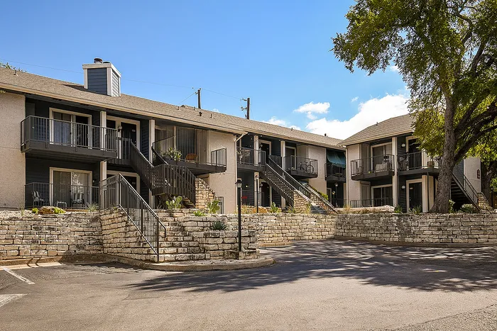 Charming apartment complex with spacious balconies, nestled among stone landscaping and lush greenery under a clear blue sky.