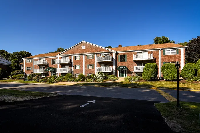 Charming brick apartment building with balconies, surrounded by lush greenery under a clear blue sky.