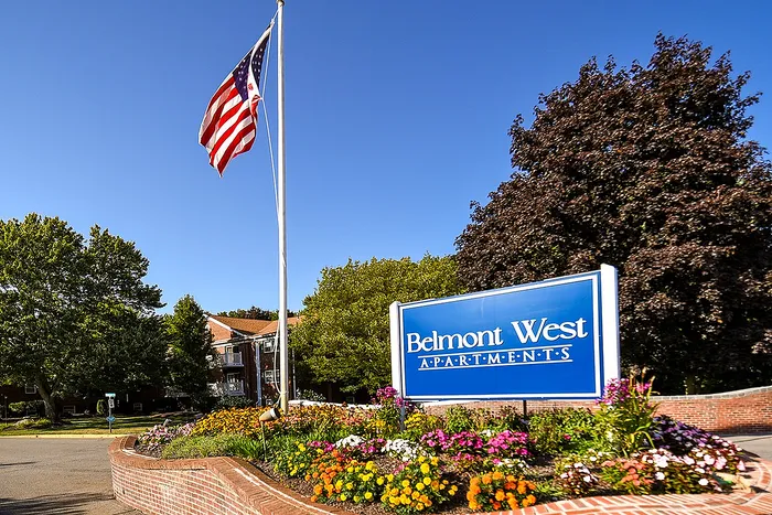 Charming entrance to Belmont West Apartments featuring a vibrant flowerbed, American flag, and welcoming community sign under a clear blue sky.