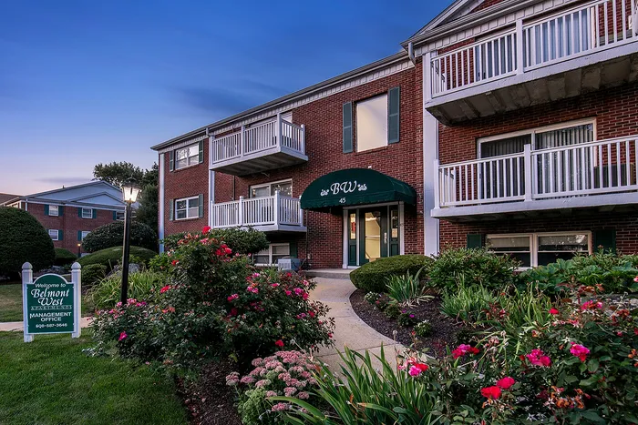 Charming brick apartment building surrounded by lush gardens and vibrant flowers, featuring inviting balconies and a welcoming entrance.