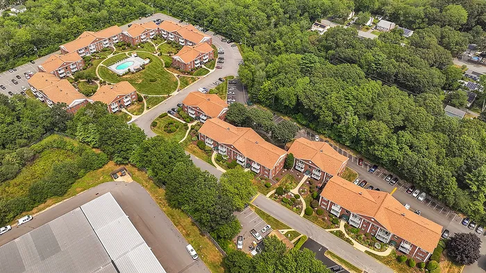 Aerial view of a vibrant apartment complex surrounded by lush greenery, featuring charming red-roofed buildings and a central swimming pool.