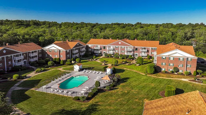 Charming apartment complex with red brick buildings and private balconies, nestled in lush greenery, featuring a pristine outdoor pool and ample sunbathing space.