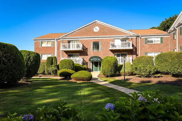 Elegant brick apartment building with charming balconies, surrounded by lush manicured gardens and vibrant hedges under a clear blue sky.