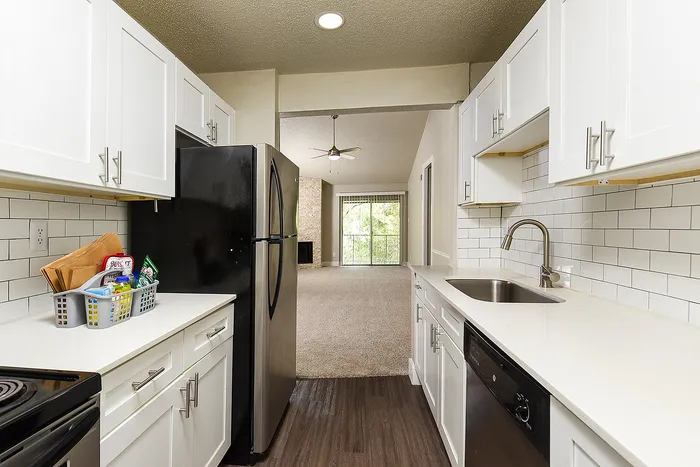 Modern kitchen with crisp white cabinetry, sleek black appliances, and elegant backsplash, leading to a spacious living area with natural light.