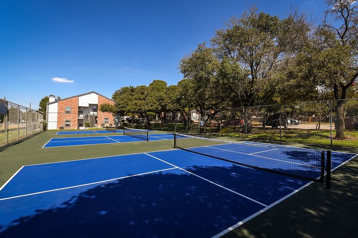 Bright pickle ball courts with vibrant blue surfaces surrounded by lush greenery and charming residential buildings under a clear blue sky.