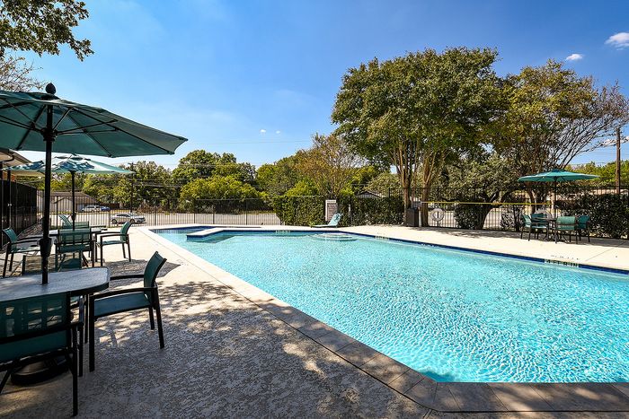 Inviting outdoor pool area with sun-kissed loungers, shaded tables, and vibrant umbrellas surrounded by lush greenery under a clear blue sky.