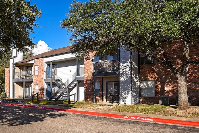 Charming apartment complex with brick facade, lush greenery, and ample parking space under a bright blue sky.