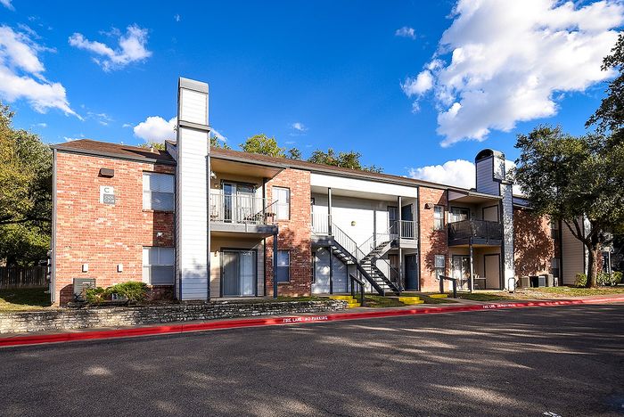Charming two-story apartment complex with red brick exterior, balconies, and lush trees under a bright blue sky.