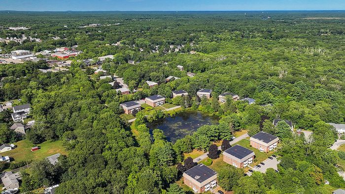 Aerial view of serene apartment complex nestled in lush greenery, featuring multiple buildings around a picturesque pond.