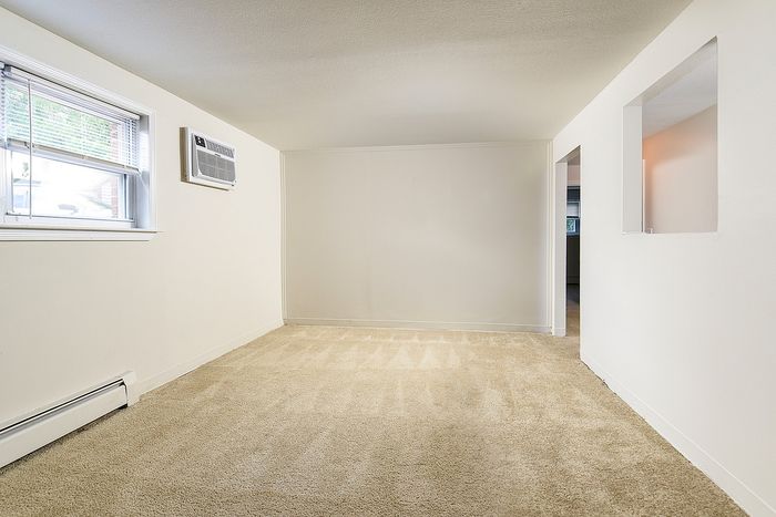 Bright and airy living space with plush carpet, large window, and a built-in air conditioning unit, offering comfort and style.