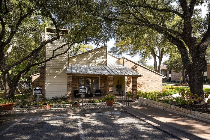 Charming brick apartment surrounded by lush green trees, featuring welcoming front porch and dedicated parking.