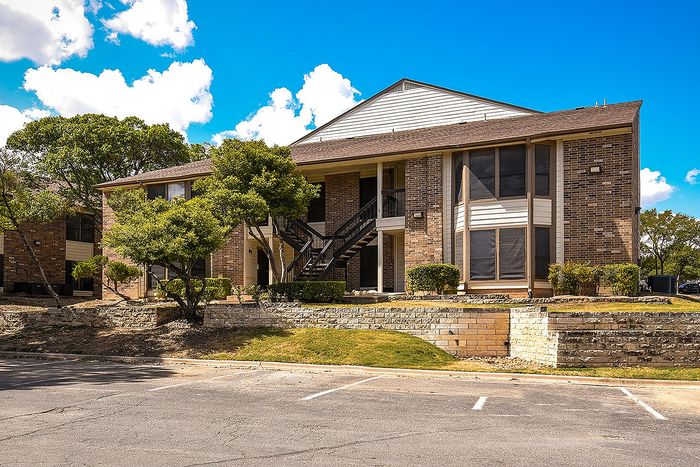 Charming brick apartment building with large windows, surrounded by lush greenery and bright blue skies.