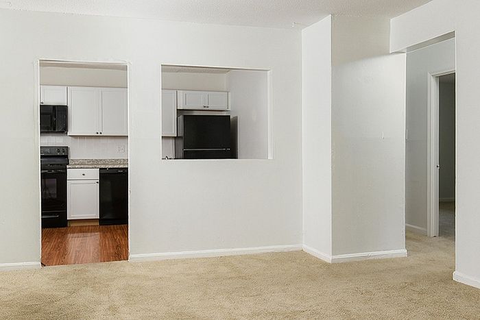 Inviting open-plan apartment showcasing a cozy carpeted living area with a view into a modern kitchen with sleek black appliances and elegant white cabinetry.