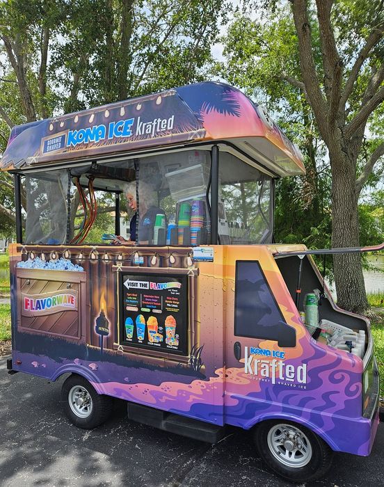 Vibrant Kona Ice truck offering gourmet shaved ice with a tropical theme, parked under lush trees.