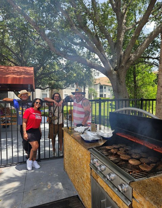 Vibrant outdoor barbecue area with a modern grill, surrounded by smiling people enjoying a sunny day under lush trees.