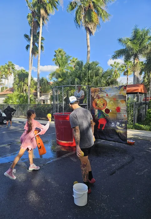 Vibrant summer festival scene with a young girl throwing a ball at a dunk tank game, set against a backdrop of tropical palm trees and a cheerful mural.