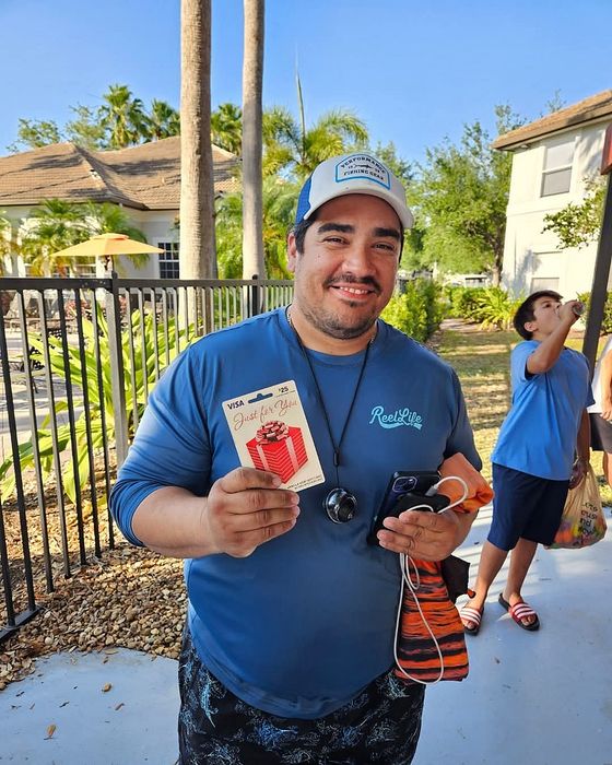 Smiling man outdoors holding a gift card, showcasing a relaxed and sunny atmosphere with tropical plants and residential buildings in the background.