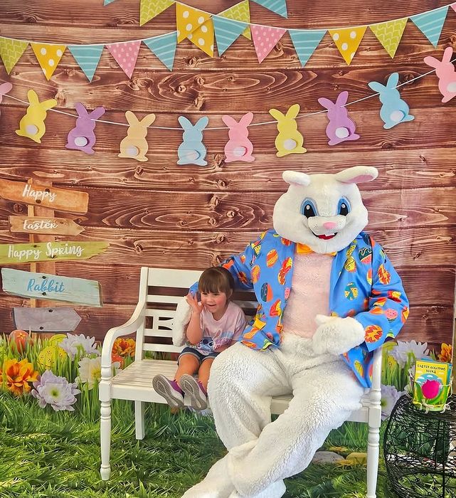 Vibrant Easter-themed setup with a cheerful bunny mascot in a colorful jacket sitting on a bench with a smiling child, surrounded by festive decorations and spring flowers.