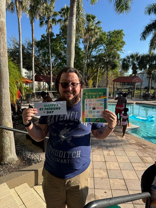 Cheerful man celebrating St. Patrick's Day by the pool under sunny skies, holding festive signs and surrounded by palm trees.