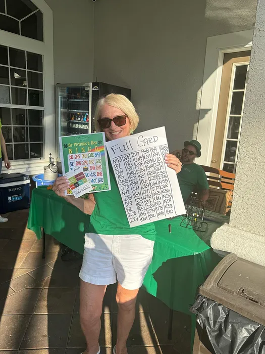 Joyful woman in green celebrating a bingo victory, holding a completed card and smiling in the bright sunlight.
