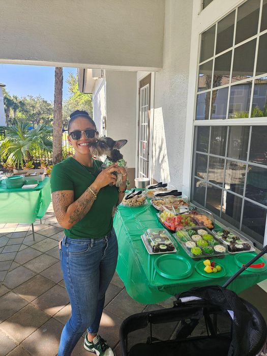 Joyful woman holding a small dog at an outdoor gathering with a vibrant food spread on green tablecloth.