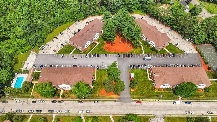 Aerial view of a lush apartment complex featuring neatly arranged buildings, ample parking, vibrant landscaping, a large swimming pool, and a tennis court, all bordered by dense, green forest.