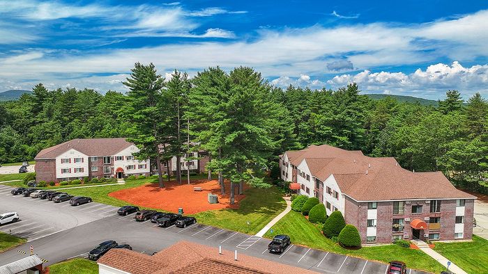 Charming apartment complex surrounded by lush greenery, offering ample parking and serene wooded views under a bright blue sky.