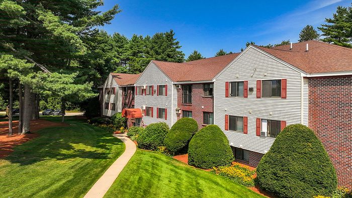 Charming apartment building with red brick accents, surrounded by lush green lawns and tall trees, offering a serene and inviting environment.