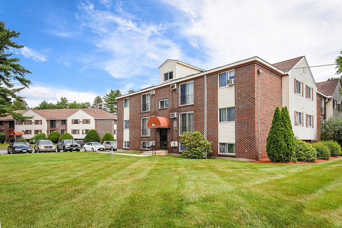 Charming brick apartment building with lush green lawn, ample parking, and picturesque surroundings under a bright blue sky.