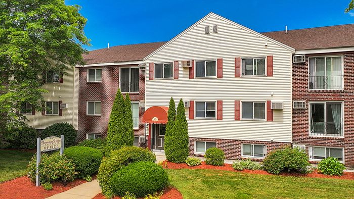 Charming apartment building with red brick and cream siding, surrounded by lush greenery and manicured shrubs.