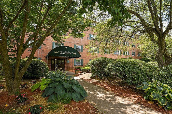 Inviting apartment building entrance surrounded by lush greenery and mature trees, offering a serene and welcoming atmosphere.