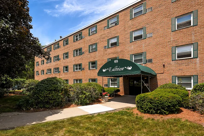Charming brick apartment building with lush greenery and inviting entrance under a green canopy at 65 Eastview.
