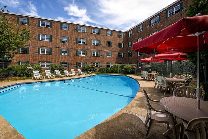 Inviting apartment complex with a large outdoor pool, surrounded by comfortable lounge chairs and vibrant red umbrellas, perfect for relaxation and socializing.