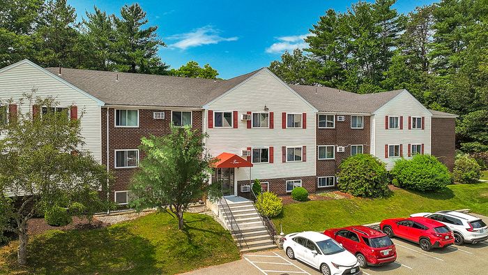 Charming apartment building surrounded by lush greenery, featuring an inviting entrance and ample parking space on a sunny day.