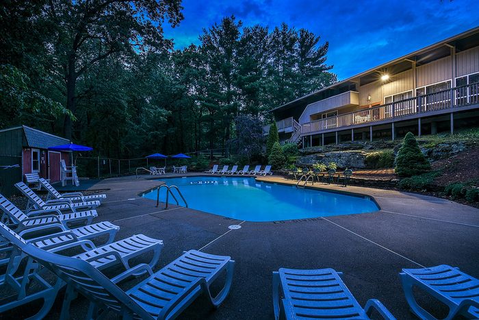 Inviting outdoor pool area with stylish lounge chairs and vibrant blue umbrellas, surrounded by lush greenery and a cozy building with warm lighting, perfect for relaxation and enjoyment.