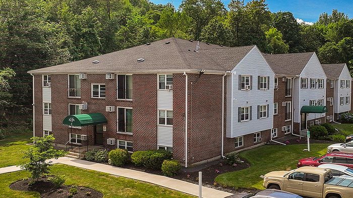 Charming red brick apartment complex with lush greenery, ample parking, and a bright, sunny day backdrop.