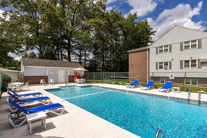 Inviting outdoor pool area with vibrant blue lounge chairs, surrounded by lush greenery and a charming brick building backdrop.