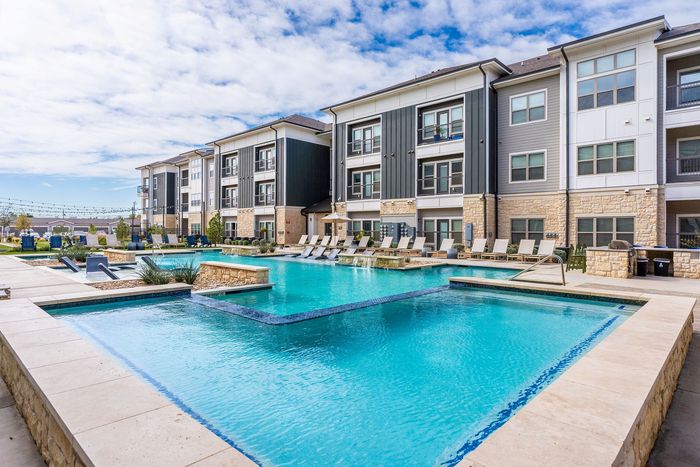 Luxurious apartment complex featuring a sparkling pool, chic poolside seating, and modern architecture under a bright blue sky.