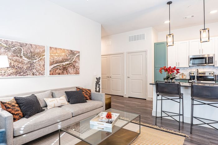 Modern and inviting open-concept living room with a cozy beige sofa, stylish throw pillows, and a sleek glass coffee table, complemented by a contemporary kitchen with elegant pendant lighting and vibrant decor.