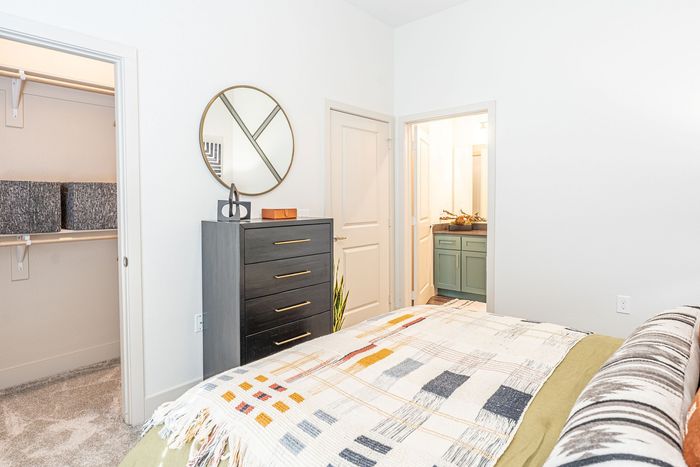 Bright bedroom with modern decor, featuring a patterned bed, sleek dresser with a stylish mirror, and an adjacent bathroom with elegant accents.