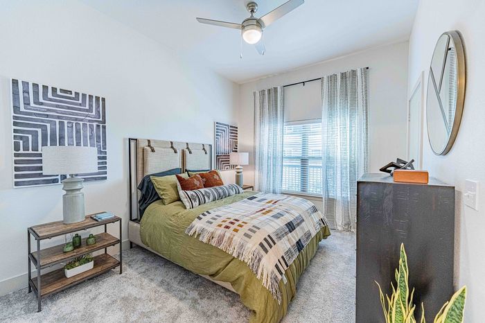 Inviting bedroom with a stylish beige and green decor, featuring a cozy bed with patterned cushions, large window with sheer curtains, modern ceiling fan, and tasteful wall art.