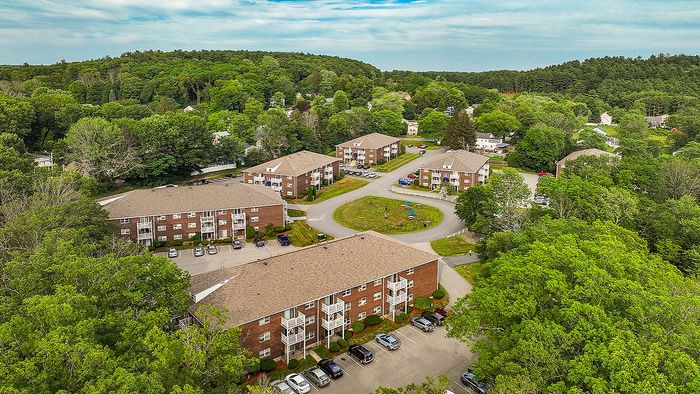 Scenic aerial view of a charming apartment complex surrounded by lush greenery, featuring multiple well-maintained brick buildings and ample parking space.