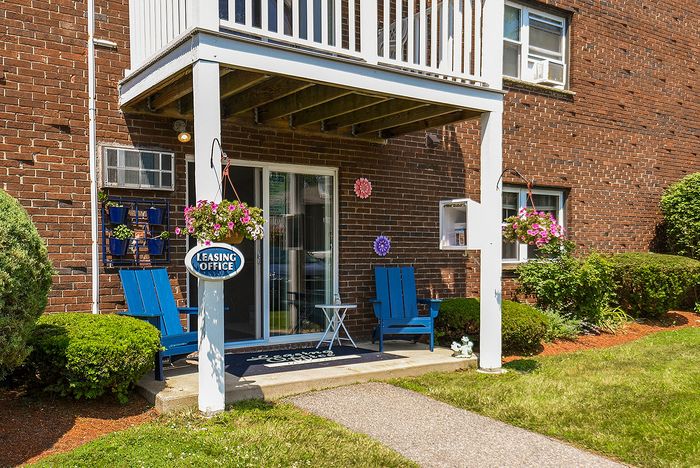 Charming apartment leasing office entrance with vibrant flowers, blue Adirondack chairs, and inviting decor.