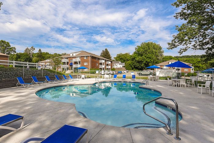 Inviting outdoor pool area with blue lounge chairs, surrounded by lush greenery and modern apartment buildings under a clear sky.