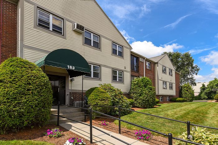 Charming apartment building with green awning, lush landscaping, and inviting entryway under a clear blue sky.
