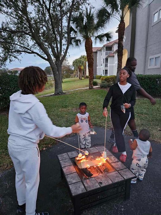 Family enjoying a cozy bonfire evening roasting marshmallows near modern apartments, surrounded by lush greenery and towering palm trees.