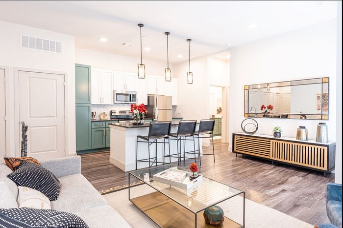Modern open-concept living space with elegant kitchen, featuring sleek cabinetry and a stylish breakfast bar with pendant lighting. Cozy seating area with a plush sofa, complemented by contemporary decor and a large mirror enhancing the room's spacious feel.